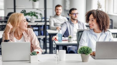 İş adamları sağlık ve sosyal mesafeyle ilgilenirler. İş yerindeki gülümseyen Avrupalı ve Afrikalı Amerikalı kadın bilgisayarları, bitkileri ve antiseptikleri kilitlendikten sonra ofiste