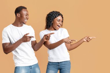 Cheerful african-american couple smiling and cheerfully pointing away at empty space, yellow studio background. Happy young black man and woman aiming at advertisement, panorama with copy space