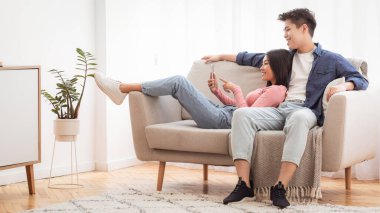 Akıllı Telefon Uygulaması Kullanan Asyalı Çift Minimalist Oturma Odasında Haftasonu Oturuyor. Telefon kullanıcıları interneti tarıyor. Gadget Yaşam Tarzı, Mobil Uygulamalar. Boşluğu Kopyala