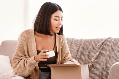 Güzellik kutusu. Meraklı Asyalı kadın paketini almış, paketleri açmış ve kozmetik ürünlerle dolu karton pakete bakmış, elinde nemlendirici krem tutuyormuş. İnternetten alışveriş yapan kadın
