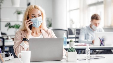 Covid-19 karantinasından sonra işine dön. Bin yıllık koruyucu maskeli bir kadın iş yerindeki bilgisayarında modern ofis içi antiseptiklerle telefonla konuşuyor, boş yer var.
