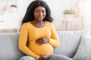 Evdeki kanepede oturan ve kameraya gülümseyen güzel siyah hamile kadın, fotokopi alanı. Neşeli Afro-Amerikalı kadın oturma odasında dinleniyor, koca karnına dokunuyor.