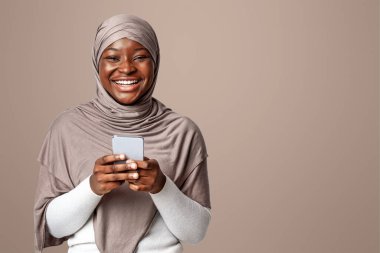 Neşeli siyahi kadın tesettürde akıllı telefon tutuyor ve kameraya gülüyor, mobil iletişimin ve online alışverişin keyfini çıkarıyor, yeni uygulamalara bakıyor, ışık stüdyosunun arka planında duruyor, Space 'i kopyalıyor.