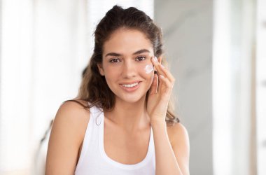 Yüzünde nemlendirici ile kameraya gülümseyen yanağa yüz kremi sürerken modern banyoda duran çekici kadının portresi. Cilt Kozmetik Ürünleri Konsepti