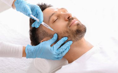 Sakallı, orta yaşlı bir adam güzellik salonunda güzellik iğnesi yaptırıyor. Kozmetik uzmanı klinikteki yakışıklı adam için yaşlanma önleyici prosedür uygulaması yapan mavi lastik eldivenleri teslim etti. Yüz bakımı, erkek kozmetoloji konsepti.