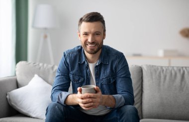 Rahat, orta yaşlı, rahat giyimli bir adam elinde bir kupa çayla kanepede oturuyor, beyaz sakallı bir adam evdeki rahat oturma odasında kahve içiyor, kameraya gülümsüyor, uzay fotokopisi çekiyor.