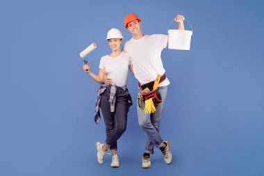 Mavi stüdyo arka planında boya kovası ve paten fırçasıyla ayakta duran mutlu ressamlar ev tadilatına hazırlar.