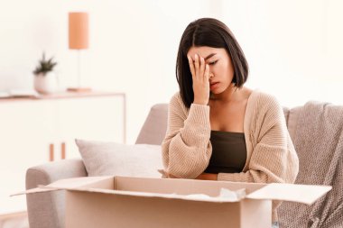 Yanlış paket. Mutsuz, hayal kırıklığına uğramış Asyalı kadın, kötü teslimat servisiyle birlikte üzgün kutusunu açıyor. Oturma odasındaki kanepede oturuyor. Sevkiyat hatası, kopyalama alanı. Avuç içiyle yüzünü kapatan kadın
