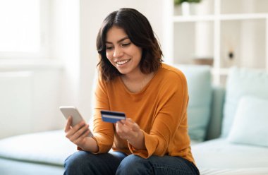 E-Ticaret Konsepti. Orta Doğulu genç bir kadın akıllı telefon ve kredi kartı kullanıyor evde koltukta otururken gülümseyen bir Arap kadın internet üzerinden ödemelerini yapıyor cep telefonunda uygulamalar, kopyalamalar yapıyor.