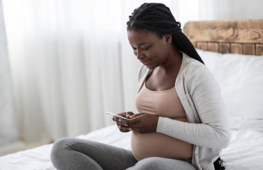 Gülümseyen Afrikalı Amerikalı kadın akıllı telefonuyla evde dinleniyor, gelecekteki anne yatakta oturuyor, arkadaşlarıyla mesajlaşıyor ya da internete takılıyor, hamilelik bloğunda bilgi okuyor, boşluğu kopyalıyor.