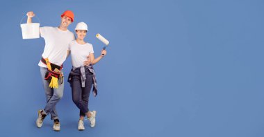 Mavi stüdyo arka planında boya kovası ve paten fırçasıyla ayakta duran mutlu ressamlar ev tadilatına hazırlar.