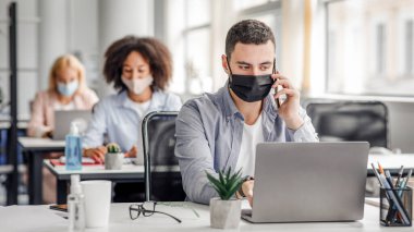 Karantinadan sonra işe dönmek için telefonla müşteri danışmanlığı. Milenyum maskeli ve akıllı telefonlu bir adam modern ofisin iç kısımlarındaki bilgisayarlara bakar.