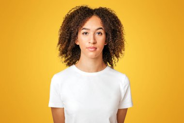 Ciddi, sakin, katı duygular. Duygusuz, duygusuz, beyaz tişörtlü, kıvırcık saçlı Afro-Amerikalı kadın ellerini arkada tutuyor ve beyaz arka planda izole bir şekilde kameraya bakıyor.