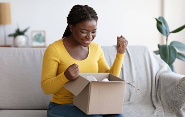 Mutlu siyahi kadın karton kutuları açıyor, başarılı alışverişe duygusal tepki veriyor, büyük alışverişlerle tatmin oluyor, heyecandan yumruklarını sıkıyor, oturma odasında kanepede oturuyor.