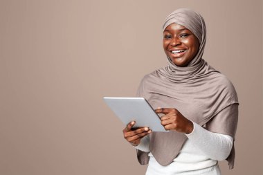 tesettürde gülümseyen siyah kadın dijital tablet kullanıyor, internetten alışveriş yapıyor veya sosyal ağlara göz atıyor, başörtülü Müslüman kadın modern teknolojilerin tadını çıkarıyor, ışık stüdyosunun arkasında duruyor, boşluğu kopyalıyor.