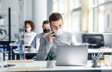 Müşteri ile uzaktan sosyal ve çevrimiçi çalışma. Korumalı maskeli adam, iş yerindeki akıllı telefonuyla konuşuyor. Coronavirus sırasında dizüstü bilgisayarı ve cam tahtasıyla oturuyor. Boşluğu kopyala.