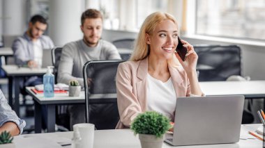 Teknolojiyle uzaktan yakından ilgilenen bir müşteriyle modern çalışma. Gülümseyen iş kadını akıllı telefondan konuşuyor ve pencereden dışarı bakıyor, işyerindeki iş yerinde bilgisayarda işçiler ile birlikte yazıyor, boş.