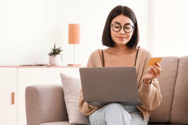 Çevrimiçi Alışveriş Konsepti. Gözlüklü gülümseyen Asyalı kadın internetten sipariş veriyor, dizüstü bilgisayar kullanıyor, elinde tutuyor ve kredi kartını gösteriyor. Bilgisayarla tatmin olmuş, koltukta oturan, boş alan.