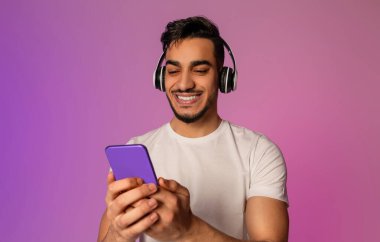 Portrait of cool young Arab man in wireless headphones using smartphone in neon light. Millennial middle Eastern guy checking new mobile app, listening to music, watching video, chatting online