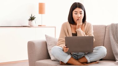 Bir sorun var. Şaşırmış ve şok olmuş Asyalı kadın bilgisayarda inanılmaz ve beklenmedik bir şey izliyor, evdeki kanepede oturuyor, boşluğu kopyalıyor. Laptop kullanan ağzı açık genç bir kadın.