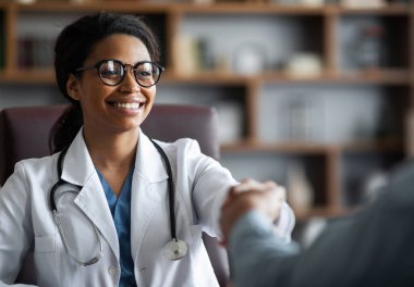 Closeup of friendly young african american lady doctor smiling and shaking male patient hand, cheerful black woman therapist greeting visitor at her clinic, outstretching hand, copy space