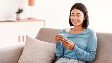 Hafta sonu eğlencesi. Akıllı telefon tutan, mesaj yazan, evdeki koltukta oturan Asyalı genç bayanın portresi. İletişim Konsepti. Mutlu kadın internette sörf yapıyor, komik videolar izliyor.