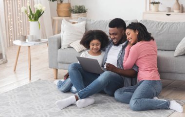Birlikte tatil yapmayı planlıyoruz. Siyah baba, anne ve küçük kız evde dizüstü bilgisayar kullanıyor, oturma odasında yerde oturuyor, boşluğu kopyalıyor.