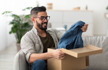 Tatmin olmuş Arap erkek müşteri karton kutudan yeni kıyafetler çıkarıyor, evdeki teslimattan memnun. Milenyum Doğulu adam karton paketleri açıyor, çevrimiçi mağaza siparişi alıyor, kapalı alanda.