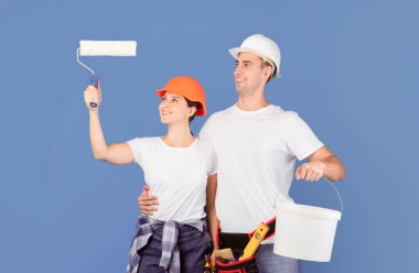 Ev Yenileme Servisi. Hardhat şapkalı mutlu çift ellerinde boya silindiri ve kova, mavi arka planda duran, duvarları boyamaya ve onarmaya hazır olumlu erkek ve kadın,