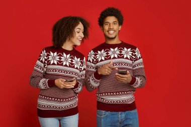 Mobil uygulamalarda yeni yıl ve Xmas promosyonları ve indirimleri. Mutlu Afro-Amerikan erkeği akıllı telefonu işaret eder ve kameraya bakar, bayan onun cihazına bakar, kırmızı arka planda izole edilmiş, stüdyo çekimi