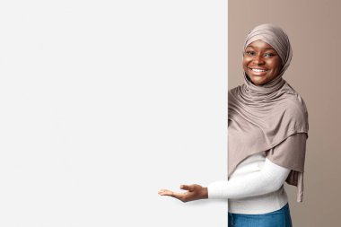 İlginç bir teklif. Beyaz reklam panosunu işaret eden tesettürlü mutlu siyahi kadın, İslami bayan, metin ya da tasarımın için fotokopi alanı gösteriyor, açık arka planda duruyor.