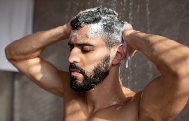 Kas sakallı, saç yıkayan, erkek şampuanı kullanan, köpüklü duş alan ve modern banyoda sıcak suyun altında duran adamın yakın plan portresi. Erkek Güzelliği Rutini ve Hijyen Konsepti