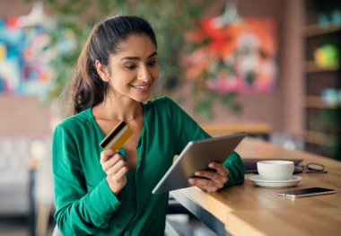 Neşeli Hintli kadın bankacılığı online, kafede dijital tablet ve kredi kartı kullanıyor, genç kadın internetten kıyafet veya kozmetik alıyor, alışveriş mobil uygulamasını ped üzerinde kullanıyor.