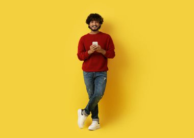Yakışıklı, kıvırcık, bin yıllık Hintli adam sarı stüdyo arka planında cep telefonuyla poz veriyor, kameraya gülümsüyor, güzel mobil uygulama kullanıyor, tam boy, kopyalama alanı kullanıyor.
