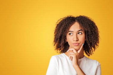 Zor seçim, teklif ya da karar vermek. Dalgın, kıvırcık saçlı, beyaz tişörtlü Amerikalı kadın parmağını çenesine bastırıyor ve sağa sola bakıyor, sarı arka planda izole edilmiş, stüdyo çekimi.