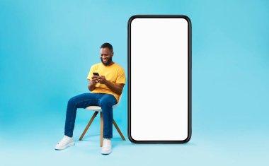 Neşeli genç siyah adam boş ekranlı büyük akıllı telefonun yanında oturuyor, mobil cihaz kullanıyor, web sitesi, reklam ya da mobil uygulama için yer açıyor, mavi stüdyo geçmişi var.