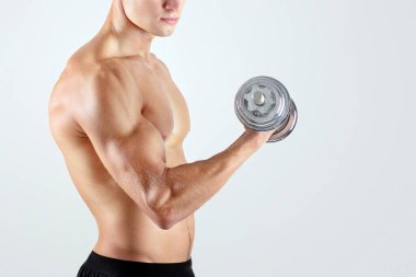 Beyaz tenli yakışıklı vücut geliştirici ağırlık kaldırıyor, hafif stüdyo arka planında dambıllarla egzersiz yapıyor. Çıplak gövdesiyle ağırlık çalışan kararlı bir milenyum sporcusu.