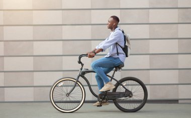 Şehir merkezindeki tuğla duvarın yanında bisiklet süren kulaklıklı zenci adamın yan görüntüsü.