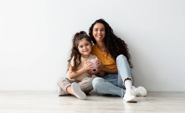 Aile Bütçesi. Neşeli Orta Doğulu Anne ve Küçük Kızı, Beyaz Duvar 'ın yanında yerde dinlenirken domuz kumbarasını tutarken, Gülümseyen Anne ve Kadın Çocuk Ekonomi ve Para Birikimlerinin keyfini çıkarırken,