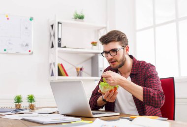 Adam modern ofis iç sağlıklı bir iş yemeği var. Genç yakışıklı işadamı çalışma yerde kase, diyet ve doğru bir konsepti yeme sebze salatası ile.