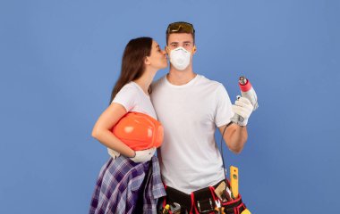 Sevgi dolu genç bir çift birlikte tamirat yapıyor, mavi arka planda inşaat aletleriyle poz veriyor, kadın elinde matkapla sert şapka tutuyor ve adamı öpüyor, daireyi yenilemekten zevk alıyor, boş alan yaratıyor.