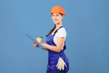 Kapaklı ve sert şapkalı mutlu tamirci kadının portresi. Tamir projesi için not alıyor, mavi stüdyonun arkasında dikilirken yenileme bütçesi planlıyor, boşluğu kopyalıyor.