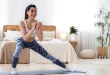 Ev aerobiği. Mutlu Asyalı kadın yatak odasındaki mindere akciğer egzersizi yapıyor, ev içi spor eğitiminin ve sağlıklı yaşam tarzının tadını çıkarıyor, şık giyiniyor, spor kıyafetleri giyiyor ve gülümsüyor.