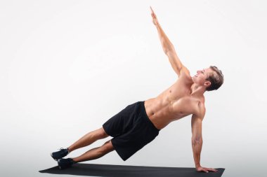 Atletik genç adamın tam boy portresi. Hafif stüdyo arkaplanı üzerine yan döşeme, fotokopi odası. Çıplak göğsü olan yakışıklı bir adam tüm vücudunu çalıştırıyor, yoga minderiyle egzersiz yapıyor.