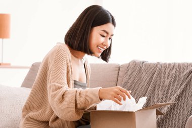 Hızlı Teslimat Servisi ve Memnun Müşteri. Gülümseyen Asyalı kadın tüketicinin portresi paket kutusunu açıp açıyor ve açıyor, mutlu kadın evdeki oturma odasında kanepede oturuyor.