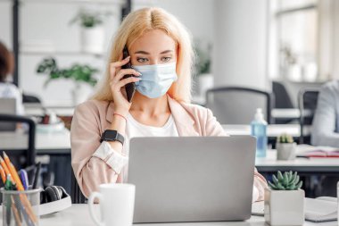 Karantinadan döndükten sonra modern ofiste çalışıyor. Koruyucu maskeli Millennial Lady telefonda konuşuyor ve masada içi antiseptik dolu bir dizüstü bilgisayara bakıyor.