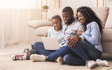 Birlikte tatil yapmayı planlıyoruz. Afrikalı baba, anne ve kız evde dizüstü bilgisayar kullanıyor, yerde oturuyor, boşluğu kopyalıyor