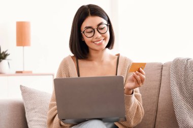 İnternetten Evden Alışveriş, Ticaret Konsepti. Gülümseyen Asyalı genç kadın evdeki koltukta oturuyor, internetten kıyafet ve ürün sipariş ediyor, kredi kartıyla ödüyor ve dizüstü bilgisayar kullanıyor.