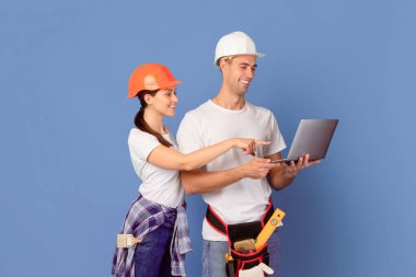 Koruyucu Miğferli Neşeli Milenyum Çifti Ev Tamiri yaparken Dizüstü bilgisayar kullanıyor, Mavi Arkaplanda Duruyor, İnşaat Malzemeleri ve Aletleri Çevrimiçi Alışveriş, Uzay Kopyalama