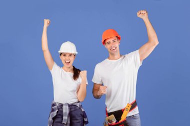 Koruyucu Miğferli Heyecanlı Genç Çift, Başarı Kutlaması. Duygusal Erkek ve Kadın, Stüdyoda Mavi Arkaplan 'ın başında dikilirken, Onarım sonuçlarının keyfini çıkarıyor.